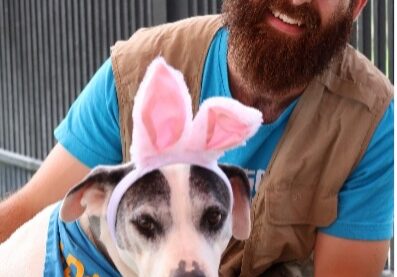 A bearded man, Nic, sits with a dog wearing bunny ears